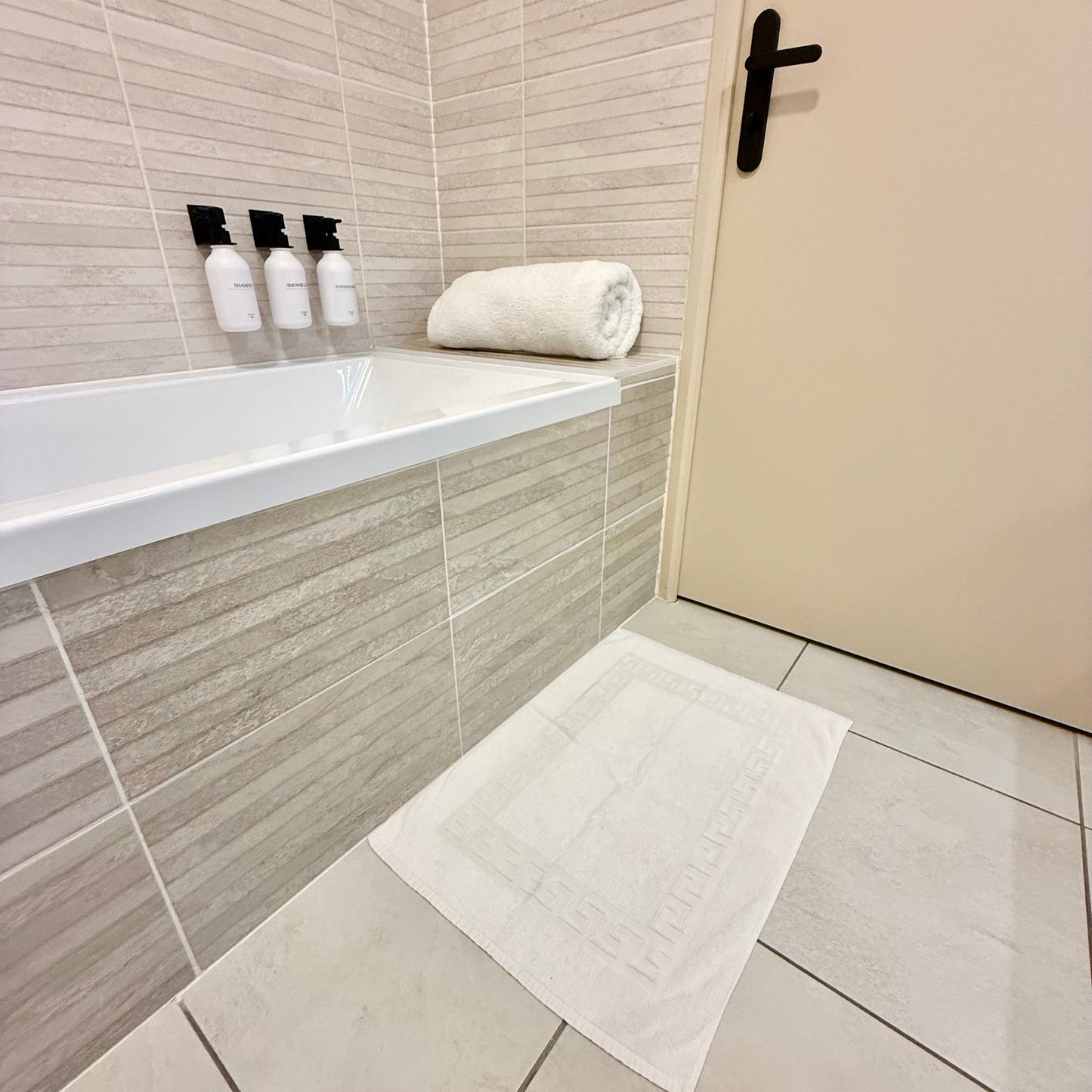 Tapis de bain blanc à motif grec en relief, posé sur un carrelage de salle de bain devant une baignoire avec produits de toilette et une serviette roulée.