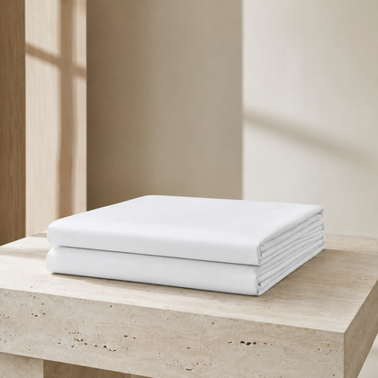 Plusieurs draps plats blancs professionnels plies sur une console, avec un lit en arriere-plan pour illustrer le linge hotelier.