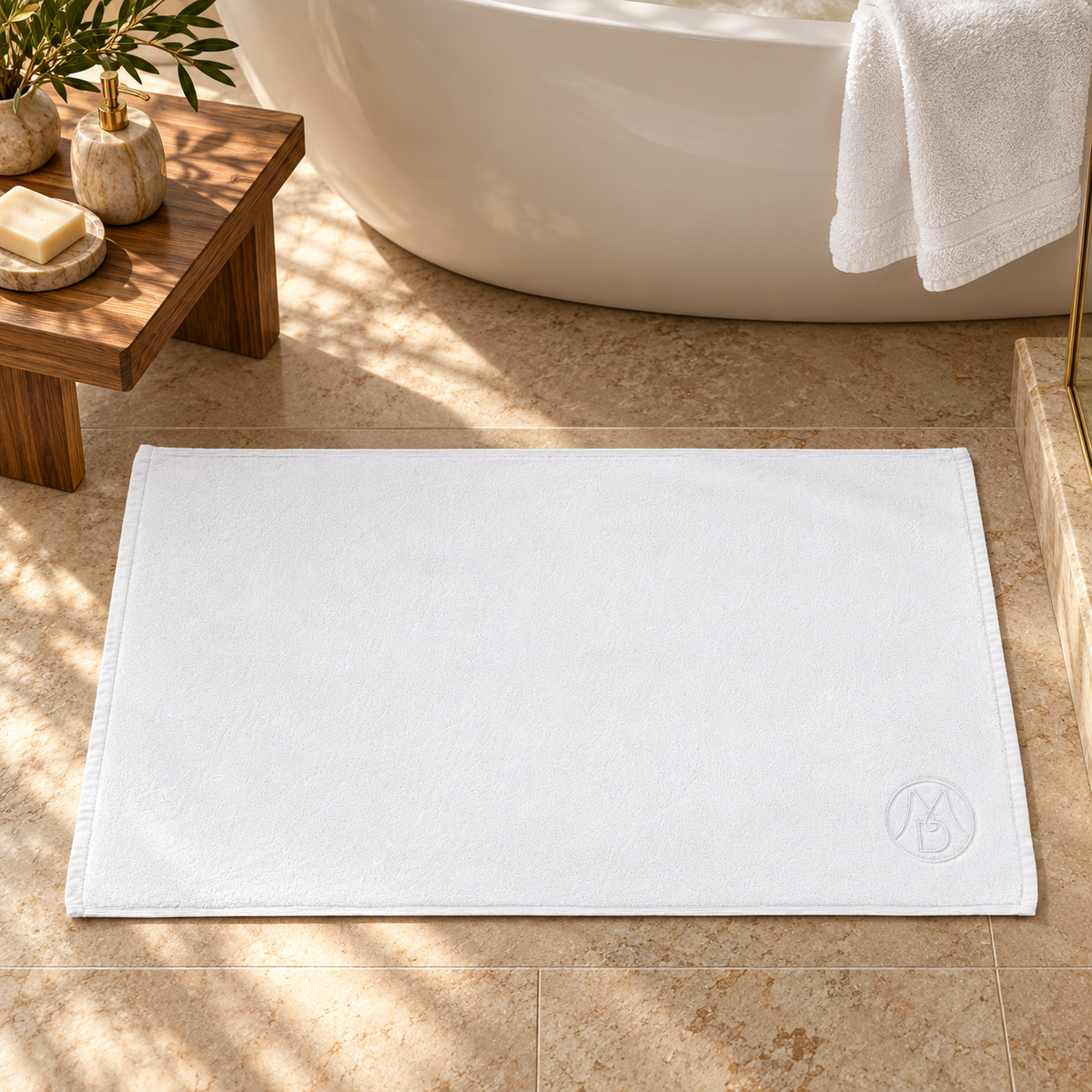 Tapis de bain hôtelier blanc en coton premium, avec monogramme brodé, posé devant une baignoire.